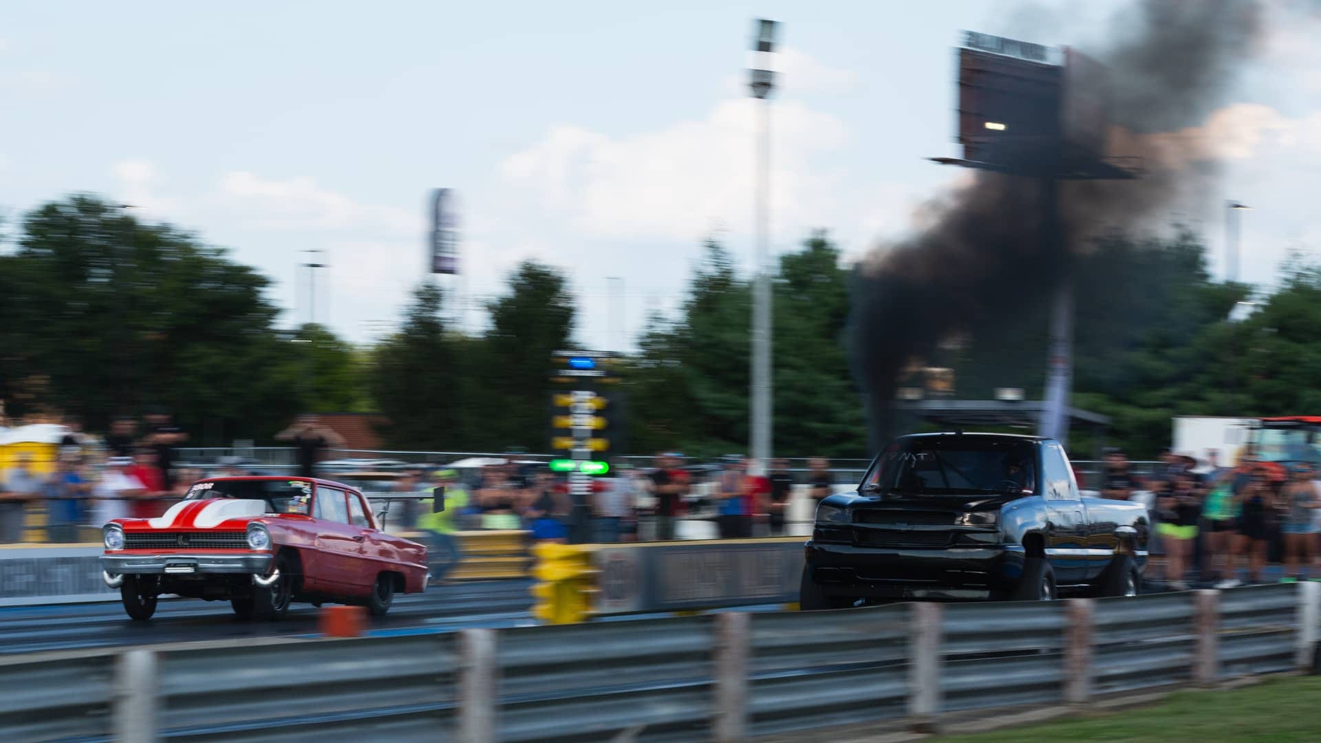 A classic car and a diesel truck launching side by side at an automotive drag event